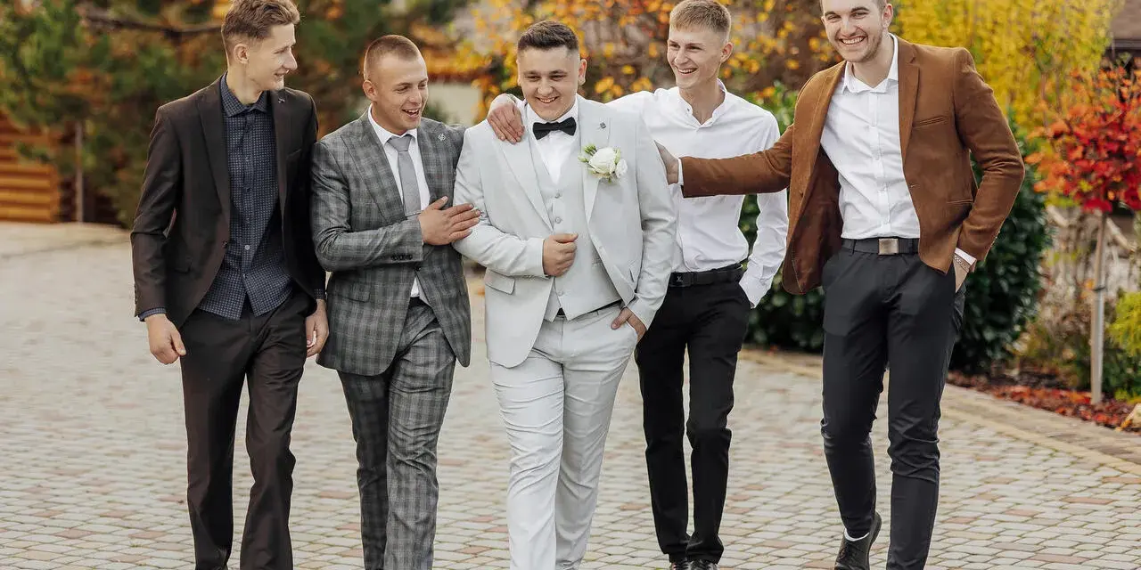 Pięciu panów w eleganckich garniturach i marynarkach, idealnie ubranych na wesele, spaceruje i rozmawia.