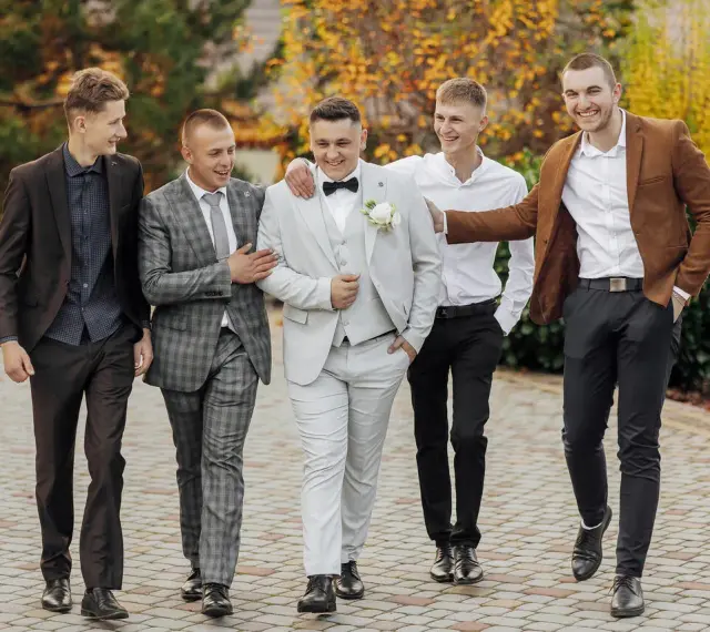 Pięciu panów w eleganckich garniturach i marynarkach, idealnie ubranych na wesele, spaceruje i rozmawia.