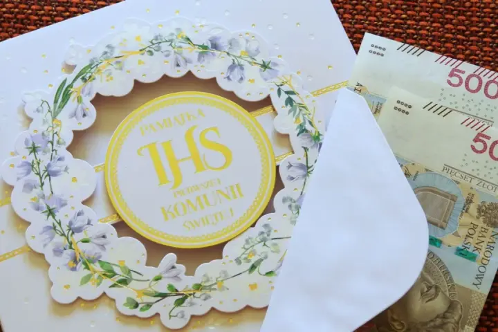 Biała kartka z motywem kwiatowym i napisem "Pamiątka Pierwszej Komunii Świętej", obok koperta z banknotami 500 zł.